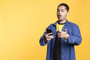 Relaxed Indian man with man bun uses smartphone browsing online apps, smiling calmly and standing against an orange studio background. Guy enjoying modern technology and wireless connection. photo