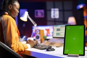 Freelancer using green screen tablet, analyzing statistical charts on laptop in living room. Close up of mockup electronic device next to remote employee checking business revenue numbers on notebook photo