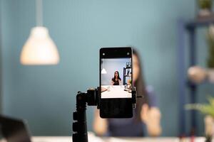 A smartphone camera captures a beauty influencer reviewing cosmetics. Mobile device screen showing asian vlogger using expressive gestures as she explains beauty products in a blue background studio. photo