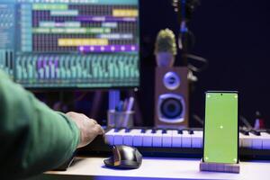 African american musician working on editing new music with a greenscreen, operating on mixing console and stereo gear in home studio. Skilled audio engineer improving sound quality. photo