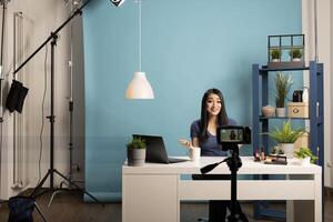 Female content creator is filming a makeup tutorial in a professional studio. Cheerful asian blogger sitting at a desk with cosmetics, speaking to the camera, with lighting and a tripod setup. photo