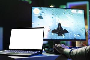 Isolated screen laptop next to man playing with friends online, shooting asteroids together in spaceship flying game. Mockup notebook next to gamer cooperating with teammates in coop videogame photo
