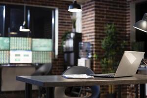 A vacant desk in modern office with a laptop and clipboard, set against a brick wall at night. Blurred computer monitors in the background create a professional and minimalist workspace. photo