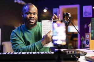 Singer producer showing musical sheet to explain notes on electronic keyboard, teaching how to play piano piano on a tutorial vlog in home studio. Performer musician presenting diatonic scale. photo