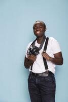 Young man in a photography studio wearing suspenders and a white t shirt. African american male individual smiles while holding his camera, exuding creativity and professionalism in a vibrant setting. photo