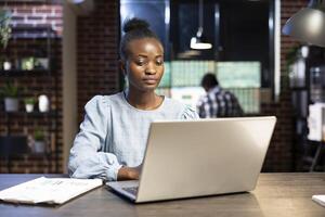 Determined female entrepreneur analyzing financial data on laptop at her desk, surrounded by trading documents. Her coworker, slightly blurred, works in the background at a multiscreen setup. photo