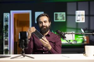 Content creator informing audience about newly launched mini soundbar. Man in studio holding high end music playing device, filming opinions with cellphone on tripod stabilizer photo