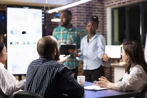 Multiethnic coworkers work efficiently in tracking progress on financial report and completing project plans. African american employees doing business presentation in late night group meeting. photo