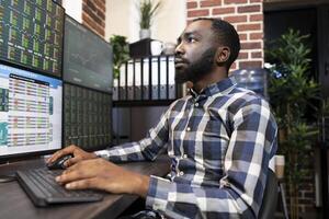 Closeup of african american stockbroker monitoring live financial charts and stock market fluctuations on computer screens. Black man utilizing his tech savvy workplace for real time trading session. photo