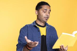 Indian male model reading a book to have fun and enjoy characters, standing with lecture against studio background. Casual guy browsing novel, enjoying leisure time and appreciating literature. photo