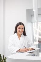 Woman in a spa robe smiles while taking notes at a wellness center reception photo