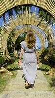 A young woman in a striped dress is seen from the back, walking along a stone pathway through a circular bamboo structure on a sunny day. video