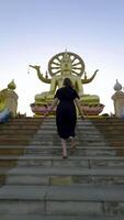 Low-angle rear view of a woman in a black dress running up a long flight of concrete stairs towards a large, golden seated Big Buddha statue on a platform under a clear, pale sky in Thailand. The video