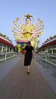 Rear view of a woman in a black dress walking towards a large, elaborate statue of Guanyin, the Goddess of Mercy, with multiple arms, adorned in gold and intricate details. The statue is located at video