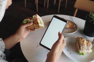 Enjoying a sandwich while browsing on a smartphone at a cafe photo