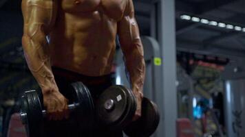 man with an impressive physique engages in bicep curls using a dumbbell in a contemporary gym setting. The environment features various gym equipment and bright lighting video
