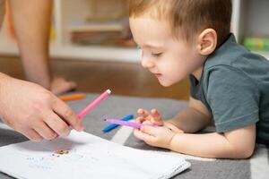A beautiful little toddler boy of two or three years old with dad draws with markers in the album in the children's room at home sitting on the floor. Spending time with children. Selective photo