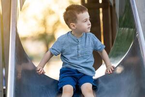 A cute caucasian toddler boy of three and a half years old going down the slide in the playground on a summer day. Active kid playing outside. Childhood concept. photo