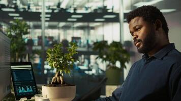 Worker in office reviewing financial graphs, analyzing business performance metrics. African american employee at work looking over data trends and strategic insights, camera B video