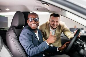 Happy african american black man in formal wear is sitting in the drivers seat of a grey sedan. New car from a dealership representative. The concept of test drive, buying an electric car or renting photo