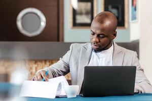 Focused african american businessman working with laptop documents in office holding papers preparing report analyzing work results, black male analyst doing paperwork at workplace using computer photo