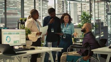 Employees and coworker with disability looking over revenue figures in inclusive office. Man in wheelchair working together with colleagues, comparing company statistical numbers, camera B video