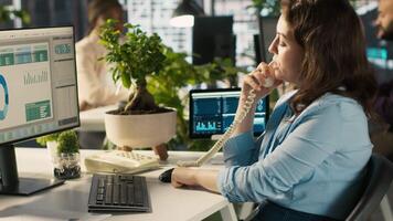 Worker in office using landline phone to talk with client while reviewing charts. Employee at work discussing with business partner during call, analyzing business performance metrics, camera A video