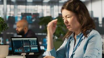 Woman suffering from migraine at work, reading medication prescription. Employee experiencing headache while reviewing financial charts on computer monitor at office desk, camera A video