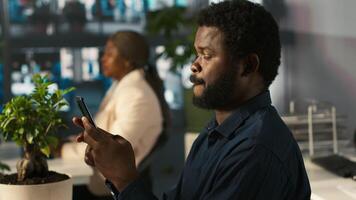 Employee in office taking break from checking documents to text friends using smartphone, relaxing. Accountant in office pausing tasks to answer messages on mobile phone, camera A video