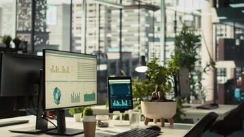 Modern workspace with analytical data dashboards displayed on devices in bright office environment. PC screen on desk with financial statistics graphs next to potted plants, zoom in shot video