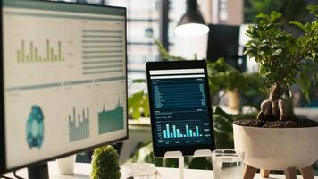 Modern workspace with key performance indicators data on display in bright office environment with potted plants. PC and tablet on desk with accounting figures displayed on screen, close up shot video