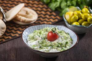 Jajeek, a refreshing cucumber yogurt salad served in a bowl with cherry tomato, pita bread and olives on wooden table side view of A classic Middle Eastern appetizer photo