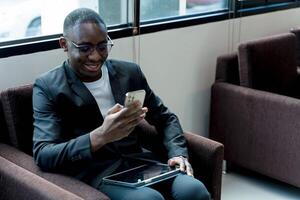 Smiling businessman using smartphone while sitting in modern lounge with tablet on lap, relaxed and engaged in communication or social media browsing photo