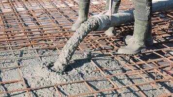 Close-up of wet concrete pouring from hose onto rebar grid with workers in rubber boots, High-detail macro of cement mix covering reinforcement for foundation construction video