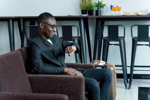 Businessman in suit checking time on wristwatch while waiting with suitcase in modern lounge, travel preparation and punctuality in professional setting photo