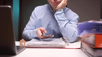 Bored office worker browsing smartphone at desk with stack of folders and miniature house video