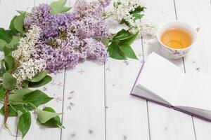 Beautiful branch of white and violet lilac flowers with opened pad, lying on the white wooden background with cup of tea, mock up perspective view with place for your text photo