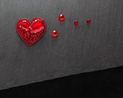 Beautiful handmade embroidered red heart brooch with crystals,lying flat on the dark gray stone rectangular background, horizontal top view with empty place for text photo