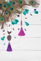 Handmade turquoise and violet bijouterie with gems, tassels and peacock feathers in boho style, lying flat on the white wooden vertical background, top view, diagonal composition. photo
