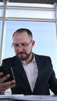 Happy businessman in office on window background. Portrait of successful entrepreneur sitting at table and reading phone message. Vertical video