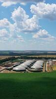 Enterprise surrounded by fields. Biogas plant among nature in bright summer day. Large storage reservoirs for biogas production. Aerial view. Vertical video