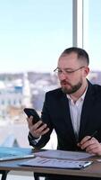 Annoyed businessman sitting at desk with a phone. Middle-aged entrepreneur in glasses using smartphone and talking to himself in office. Business problems. Vertical video
