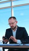 Focused businessman uses smartphone in workplace. Bearded entrepreneur in suit and glasses sitting at table and working next to large window. Vertical video