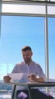 Businessman at workplace on office windows background. Handsome man in white shirt sitting at the table with a laptop and working on new project. Vertical video