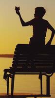Little boy is playing with a paper boat on an old bridge by the river. Silhouette of little boy sitting alone at the river on sunset background Vertical video