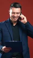 Happy businessman talking the phone. Handsome man in suit holding folder and speaking on the phone in studio. Office manager on dark red background. Vertical video