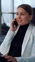 Woman having a break from work and speaking on the phone. Relaxed smiling lady sitting in bean bag chair and looking into window chatting on the smartphone. Vertical video