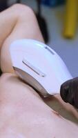Relaxed Caucasian male lies on the couch holding hands behind his head. Gloved hand of cosmetologist applies Elos laser to remove hair on male armpits. Top view close up. Vertical video
