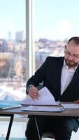 Portrait of a businessman at workplace. Bearded office worker in suit sitting at the table with a laptop on window background in the city. Vertical video