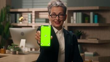 Old middle-aged European business woman showing mobile phone web smartphone green screen display chroma key copy space recommend thumb up like app good mature female employer businesswoman in office video